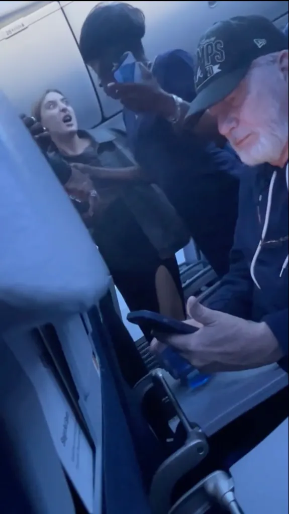 An airline passenger in a black jacket yells at an unseen person.